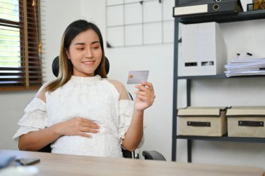 Çekici Asyalı hamile iş kadını ya da kadın patron karnına dokunuyor ve masasında otururken kredi kartı tutuyor. Bebeği için finansal yatırım planı yapmayı planlıyor..