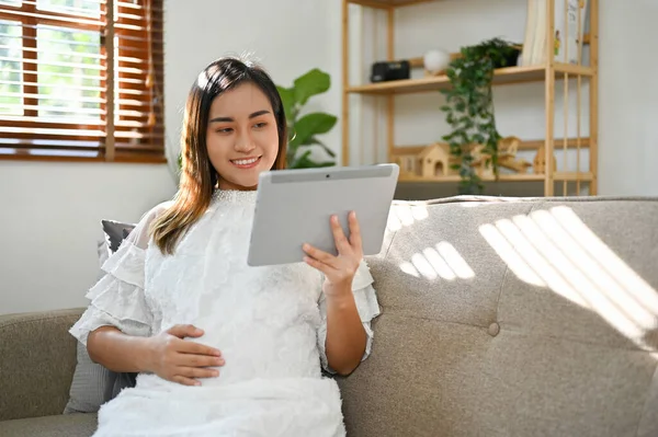 快乐的亚洲孕妇坐在沙发上 躺在客厅里 摸着肚子 在平板电脑上看婴儿护理 母性概念 — 图库照片