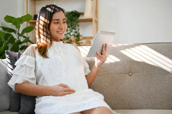 快乐而美丽的亚洲孕妇躺在客厅里 摸着肚子 在平板电脑上看婴儿护理 母性概念 — 图库照片