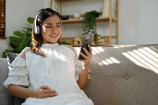 迷人的亚洲孕妇 戴着耳机 一边听音乐 一边在网上观看新生儿保育视频 一边坐在客厅的沙发上 — 图库照片