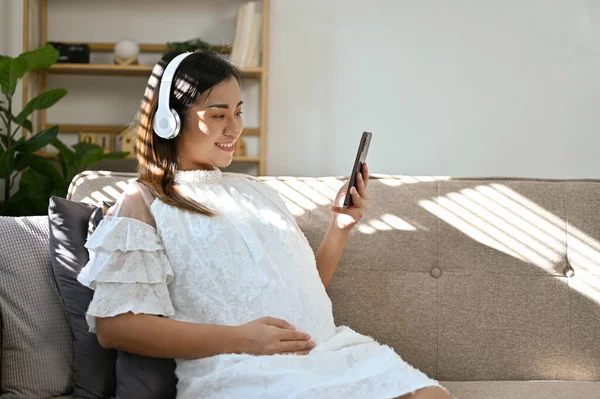 快乐而美丽的千年亚洲孕妇坐在客厅的沙发上 一边用手机 一边用耳机听音乐 — 图库照片
