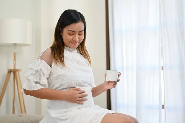 Happy and relaxed Asian pregnant woman touching her belly with love, feeling her baby in her stomach, sipping hot tea while resting in her living room. motherhood concept