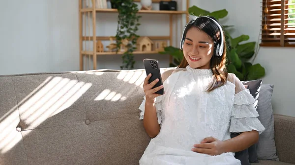 亚洲孕妇头戴耳机 在客厅里听音乐或观看网上的新生儿护理视频 这让她们感到轻松和害怕 — 图库照片
