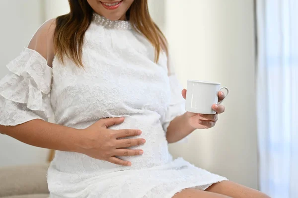 Happy Asian pregnant woman touching her belly with love, feeling her baby in her stomach, sipping hot tea while resting in her living room. cropped shot