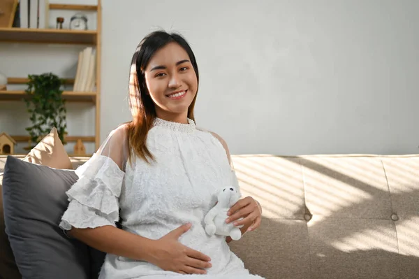 快乐而美丽的亚洲孕妇摸着她的肚子 抱着一只毛茸茸的北极熊坐在沙发上 母性概念 — 图库照片
