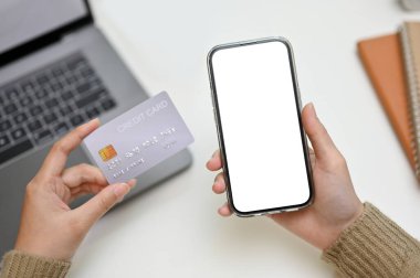 Modern ofis masasının üzerinde kredi kartı ve akıllı telefon tutan bir kadın eli. Afişinizi göstermek için telefon beyaz ekran modellemesi.