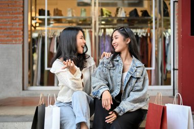 İki neşeli ve mutlu Asyalı kadın mağazanın önünde birlikte otururken konuşmaktan ve gülmekten zevk alıyorlar. Alışveriş ve alışveriş konsepti