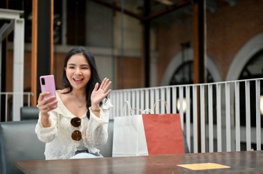 Güzel genç Asyalı kadın akıllı telefonunu kullanıyor. Alışveriş merkezinin önünde otururken arkadaşıyla görüntülü konuşuyor..