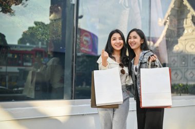 Alışveriş merkezinin önünde alışveriş torbalarıyla bekleyen iki çekici ve mutlu Asyalı genç bayan müşteri. Alışveriş ve yaşam tarzı konsepti