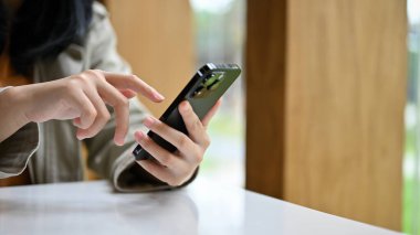 Kafe masasında oturan ve akıllı telefonunu kullanan sıradan giysili genç Asyalı kadın. dokunmatik ekran, mesaj, mobil uygulama kullanarak. kırpılmış ve yakın plan resim