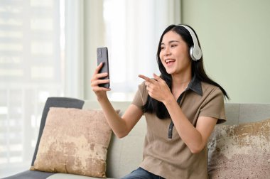 Neşeli ve güzel Asyalı kadın, günlük kıyafetler içinde, kulaklık takıyor, akıllı telefonuyla arkadaşlarıyla konuşmaktan hoşlanıyor, oturma odasında dinlenirken..
