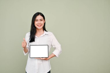 Beyaz tişörtlü güzel bir Asyalı kadın elinde dijital tablet beyaz ekran maketi ile baş parmağını kaldırıp gri arka planda duruyor. Tavsiye ya da öneri.