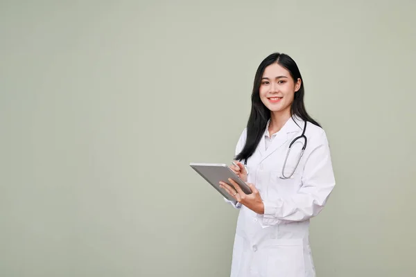 身穿白袍的美丽而专业的亚洲女医生 她的听诊器上拿着一个数码平板电脑 站在灰色的背景下 与外界隔绝 — 图库照片
