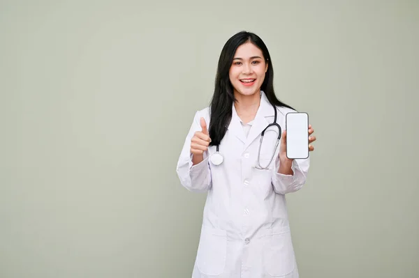 白いガウンの魅力的なアジアの女性医師は スマートフォンの白い画面のモックアップを保持し 灰色の背景に立って スタンプを示す聴診器で 素晴らしい 素晴らしい 素晴らしい — ストック写真