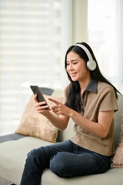 stock image Portrait, Charming and pretty young Asian woman wearing headphones, using her smartphone, listening to music while sitting on sofa in the living room.