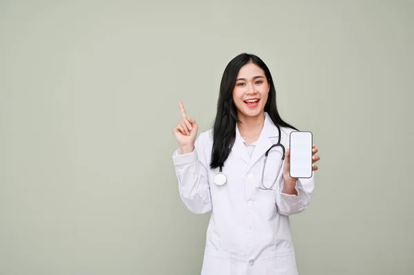 白い画面のスマートフォンを持ち 指を指して 灰色の背景に隔離された立っている制服を着た美しく笑顔の若いアジアの女性医師 健康申請 — ストック写真