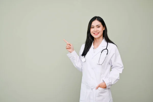 専門的かつ魅力的な若いアジアの女性医師で制服と聴診器指差し指アップ カメラを見て 孤立した灰色の背景の上に立って — ストック写真