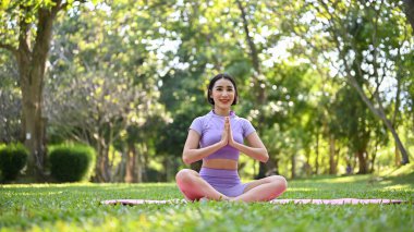 Mutlu ve rahat Asyalı kadın spor giyim kuşağında yeşil parkta meditasyon yapıyor, sakin ve nefesine odaklanmış hissediyor..