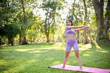 Spor kıyafetleri içinde sağlıklı ve formda genç bir Asyalı kadın. Kollarını esnetiyor, antrenmandan önce vücudunu ısıtıyor. Güzel doğa parkında spor minderi üzerinde egzersiz yapıyor..