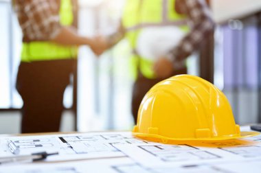 Mühendis ya da inşaat işçisi çalışma alanı. Masasında sarı bir şapka ve mavi bir iz var. Arka planda iki işçinin el sıkışması var. yakın plan resim