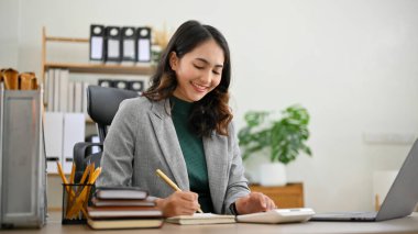 Charming and professional millennial Asian businesswoman or female manager working at her desk, taking notes on her schedule book. clipart
