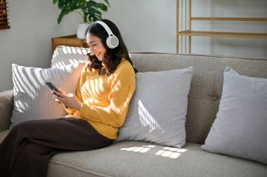 Mutlu, genç Asyalı kadın, günlük giysiler içinde hafta sonunu evinde, kanepede dinlenerek, kulaklıkla müzik dinleyerek ve akıllı telefonuyla geçiriyor..