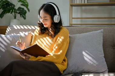 Çekici ve rahat genç Asyalı kadın günlüğünü oturma odasında yazarken kulaklıkla müzik dinliyor..