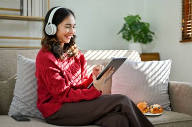 Kırmızı kazaklı neşeli ve güzel bir Asyalı kadın, kulaklık takıyor, dijital tabletini kullanıyor, oturma odasında kanepede oturuyor..