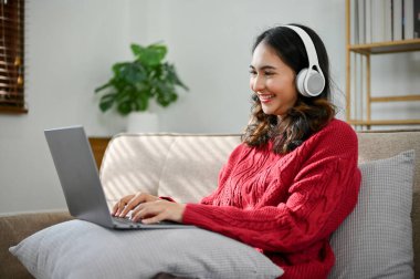 Yan görünüş, kırmızı kazaklı güzel Asyalı kadın oturma odasındaki koltukta dinleniyor, kulaklık takıyor ve taşınabilir dizüstü bilgisayarını kullanıyor..