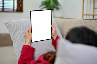 Genç Asyalı kadın oturma odasında dinleniyor, kanepede yatarken tablet ekranından bir şey izliyor. tablet beyaz ekran modeli.