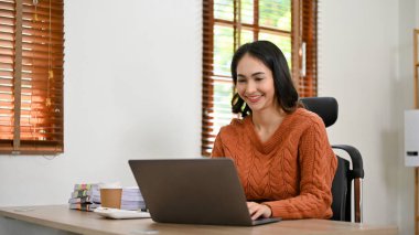 Profesyonel ve neşeli Asyalı iş kadını ya da ofis masasında çalışan günlük kıyafetlerle bayan yönetici, görevlerini yerine getirmek için dizüstü bilgisayar kullanıyor..