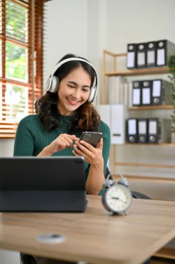 Portre, Mutlu Asyalı kadın ofis çalışanı masasında mola veriyor, kulaklıklarıyla müzik dinliyor, cep telefonunu kullanıyor..