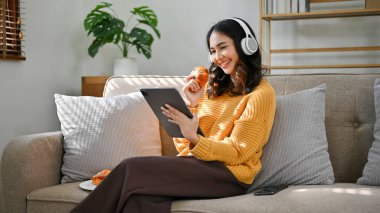 Çekici, genç Asyalı kadın kulaklık takıyor, tabletten bir şey izliyor ve oturma odasında dinlenirken lezzetli fırınlar yiyor..
