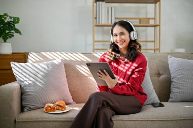 Kırmızı kazaklı çekici ve güzel bir Asyalı kadın. Kulaklık takıyor, tablet tutuyor, gülümsüyor ve oturma odasında kanepede otururken kameraya bakıyor..