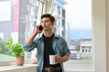 Bin yıllık yakışıklı Asyalı erkek çalışan ya da günlük giysiler içinde iş adamı pencerenin kenarında durup telefonla konuşuyor ve elinde kahve fincanı tutuyor..