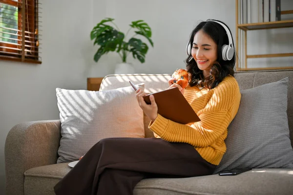 美丽的千年亚洲女性 身穿舒适的毛衣 坐在沙发上 通过耳机听音乐 一边看书 一边享受美味的面包 — 图库照片