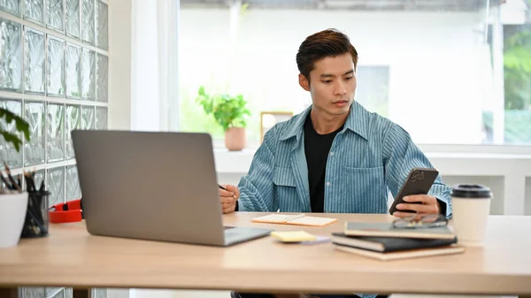 Stock image Smart young Asian businessman or male freelancer in casual outfit using his smartphone while working on his tasks at his workplace.