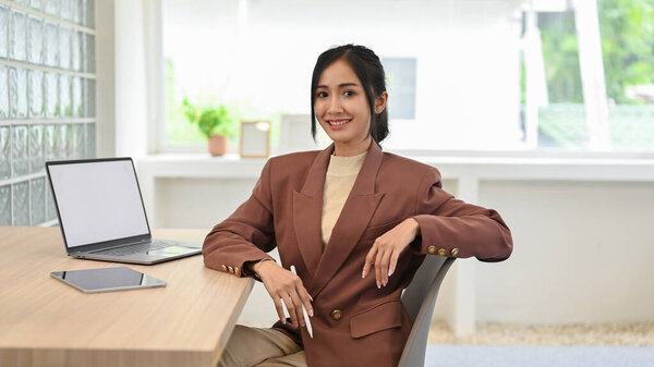 Professional and successful Asian businesswoman in casual business suit sits at her office desk, smiling and looking at camera.