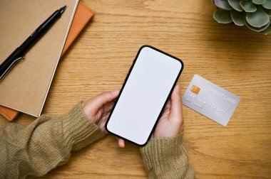 Üst manzara, beyaz ekranlı bir akıllı telefon modeli bir kadının elinde, kredi kartıyla ahşap masa arkasında..