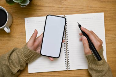 Akıllı telefon beyaz ekran maketi ve kalem tutan bir kadın el ahşap masasında çalışıyor ya da ödev yapıyor. en üstteki görünüm