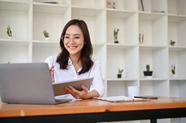 Güzel ve çekici Asyalı genç iş kadını ya da gözlüklü bayan pazarlamacı, ofis masasında çalışıyor, pazarlama projesini planlıyor ve dizüstü bilgisayar kullanıyor..