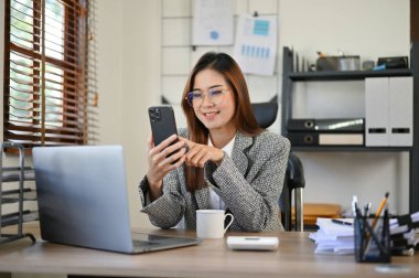 Güzel ve çekici Asyalı iş kadını gözlüklü ofis masasında oturmuş, akıllı telefonunu arkadaşlarıyla sohbet etmek için kullanıyor..