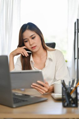 Portre, çekici ve yakışıklı Asyalı genç iş kadını ofis masasında çalışıyor, dijital tablet dokunmatik ped kullanıyor, yeni seçim kampanyasını düşünüyor..