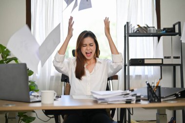 Angered, furious, crazy and mad millennial Asian businesswoman or female office worker screaming, shouting and throwing up papers at her office desk. clipart