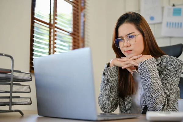 stock image Thoughtful young Asian businesswoman or female manager in eyeglasses thinking on her new project and searching her inspiration ideas on the internet, using laptop computer.