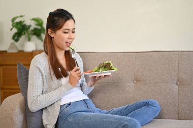 Sağlıklı ve yakışıklı Asyalı kız oturma odasında dinlenirken sağlıklı salatasını yiyor. Sağlıklı ve sağlıklı yaşam tarzı kavramı.