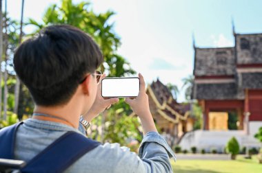 Yakışıklı genç Asyalı gezgin ya da akıllı telefonunu kullanarak güzel Kuzey Tayland tapınağının fotoğrafını çeken sırt çantalı bir gezgin. Telefon beyaz ekran modeli. Tatil konsepti. arka plan