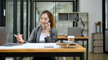 Güzel ve profesyonel Asyalı genç iş kadını ya da iş ortağıyla telefonda konuşurken ofisteki görevi üzerinde çalışıyor..