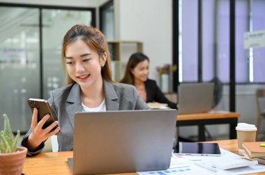 İş yerinde çalışan çekici Asyalı genç bayan ofis çalışanı masasında çalışırken akıllı telefonunu kullanıyor..