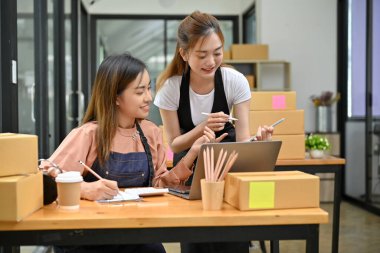 İki çekici Asyalı genç bayan arkadaş depo odasında birlikte çalışıyorlar, nakliye kutuları hazırlıyorlar ve siparişleri kontrol ediyorlar. e-ticaret iş konsepti